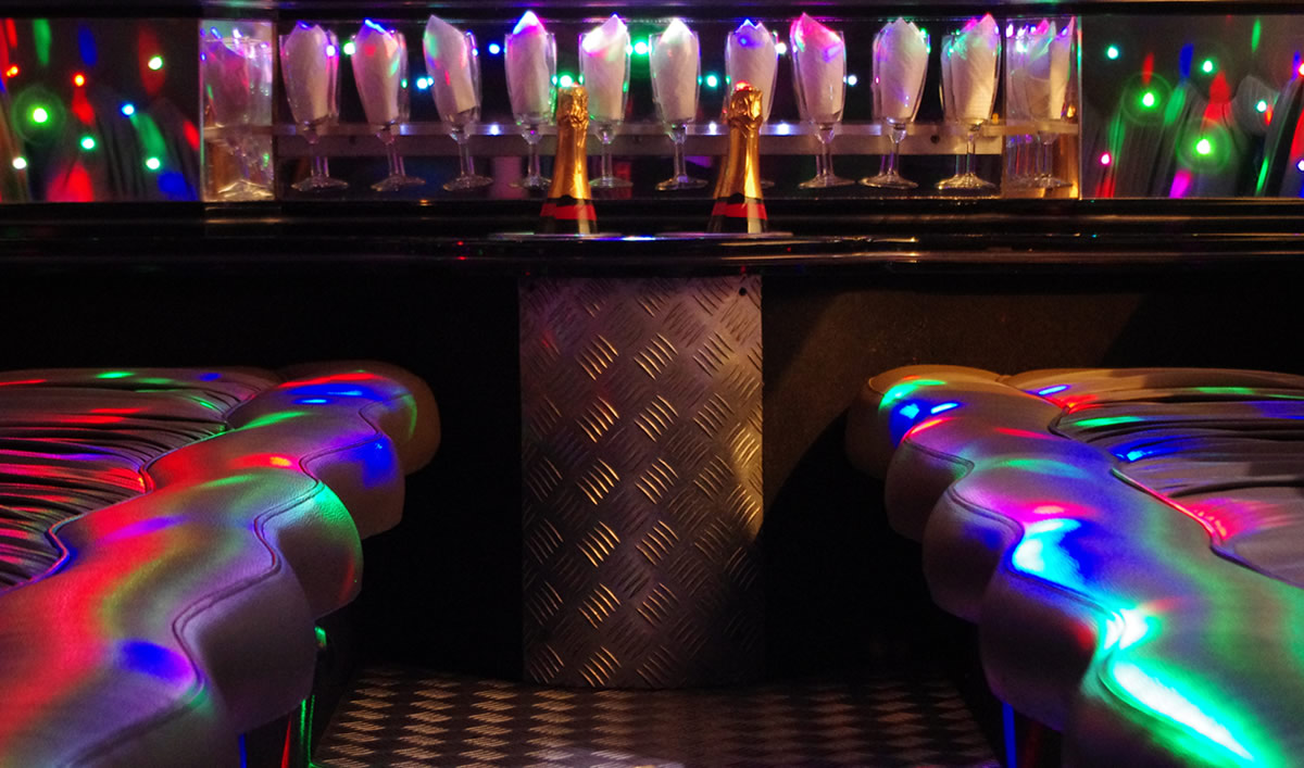 View of black 4x4 limousine interior showing seating, coloured lighting and a set of champagne flutes in the bar area